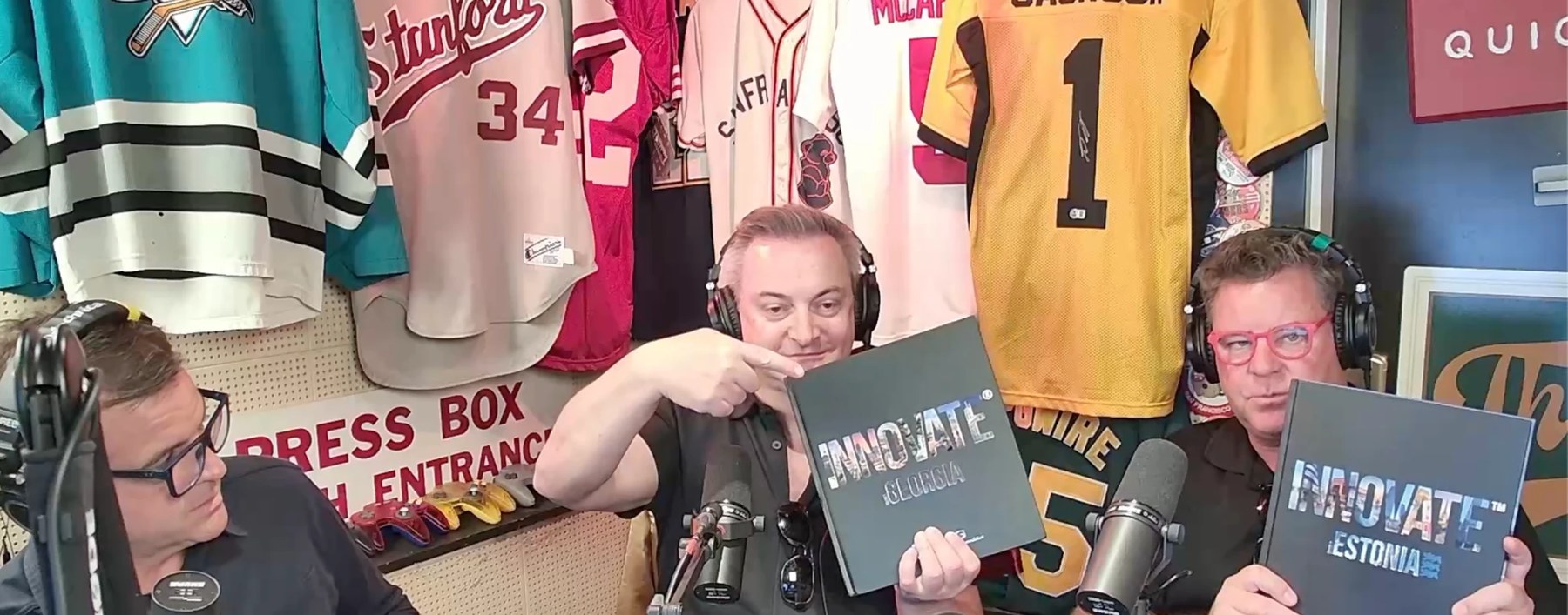 Three hosts in the podcast studio in downtown Palo Alto, surrounded by framed sports jerseys, holding up copies of INNOVATE Georgia and INNOVATE Estonia.